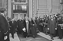 Photo en noir et blanc d'individus sortant d'un bâtiment ; au centre, une femme souriante, coiffée d'un chapeau, portant un long collier et un manteau en fourrure tient le bras d'un homme en costume trois pièces, cheveux gris-blancs, moustache grisonnante, chapeau à la main ; ils sont entourés d'autres hommes en tenue civile et de gardes républicains