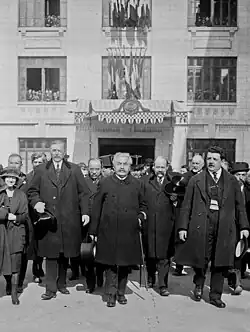 Photo en noir et blanc d’un groupe d'individus marchant dos à un bâtiment officiel ; au centre, un homme aux cheveux gris-blancs et à la moustache grise tient un chapeau dans sa main droite et une canne sous son bras gauche
