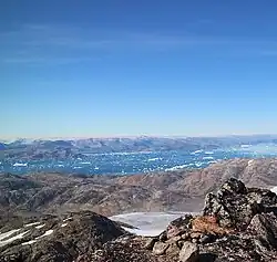 Photographie en couleurs d'un sommet montagneux comportant un cirque de glace, les eaux d'un fjord apparaissant en contrebas et en arrière-plan.