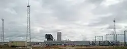 Des installations industrielles comportant plusieurs tours et structures métalliques, à droite et à gauche de l'image et des bâtiments, devant un ciel nuageux.