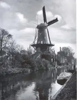 Photographie en noir et blanc d'une voie d'eau, une maison couronnée d'un moulin et entourée de végétations sur la berge de gauche, deux barques à quai sur la berge de droite.