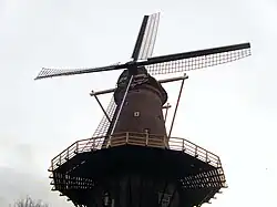 Photographie en couleurs de la partie supérieure d'un moulin à vent vue du dessous pointant vers le firmament d'un ciel nuageux.