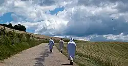 Trois moines chartreux de dos marchent sur une route goudronnée, entre deux champs fauchés, et le ciel est bleu et nuageux au troisième plan.