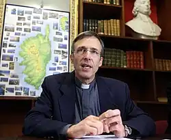 Photographie couleur d'un homme assis devant une carte murale de la Corse.