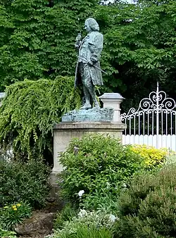 Statue en pied d'un homme en costume dix-huitième siècle, entourée de verdure.