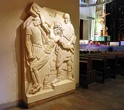 Photo de l'intérieur d'une grande église, avec à droite vue sur la nef et l'autel moderne, à gauche panneau en bas relief représentant la crucifixion