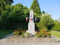 Photographie d'un monument en forme de petit obélisque au milieu d'une aire gravillonnée entourée de haies