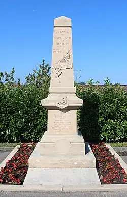 Un monument aux morts blanc en forme d'obélisque entouré par des fleurs rouges à sa base.