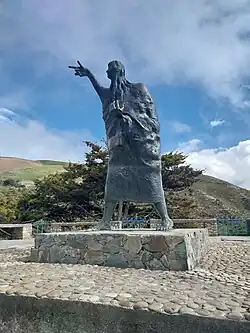 Une très grande femme en bronze moderne, debout avec de longs cheveux, désignant quelque chose de la main. Elle se trouve sur un double piédestal en pierre, devant une colline désolée.