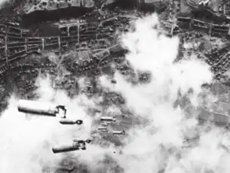 Photographie en noir et blanc prise en contre-plongée depuis un bombardier en train de déverser des bombes sur une ville.