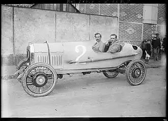 Photo en noir et blanc montrant deux hommes assis au volant d'une voiture ancienne, sans toit.