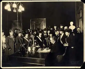 A man sits at a desk sigining a document while a large group of women watches
