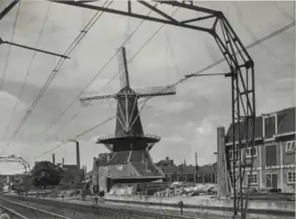 Photographie en noir et blanc d'un caténaire et des rails de chemin de fer traversant un paysage urbain, un moulin à vent et des bâtiments se dressant à droite de la ligne ferroviaire.