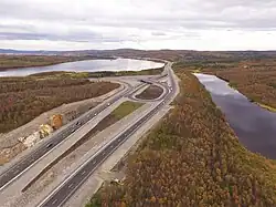 Vue aérienne d'une 2 x 2 vois allant du coin en bas à gauche de l'image au fond de l'image en disparaissant dans le paysage. Au second plan, un échangeur en trompette se dessine, avec un lac en haut à gauche et un autre lac à droite. La taïga recouvre le paysage, avec des tons automnals. En arrière-plan se distingue la silhouette d'une zone urbaine avec des collines encore derrière.