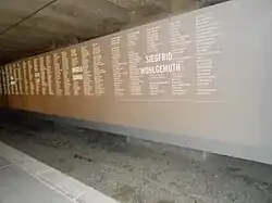 Image d'un mur gris en diagonale gravé de noms sous une toiture.