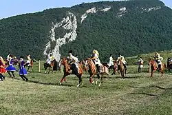 Cavaliers au galop et danseurs sur fond de paysage montagneux.