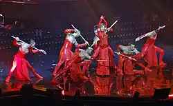 un groupe de six danseurs torses nus vêtus de rouge portant un baton sur les épaules autour de Mylène Farmer