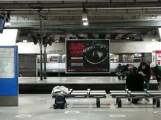 sur un quai de métro, une rame est en arrière fond, au centre l'affiche du spectacle Nermore où Mylène Farmer apparaît au milieu d'un oeil noir. Des passages attendent le métro à droite de la photo.