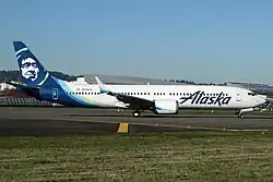 Photo de l'avion posé au sol. Un 737 blanc, avec inscrit "Alaska" sur l'avant, et un visage inuit en tons de bleu sur l'empennage.