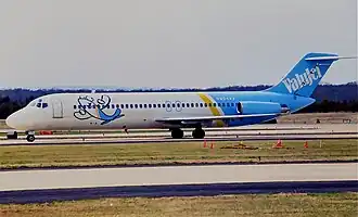 Photo du côté gauche d'un avion de ligne aux couleurs blanches à l'avant et bleues à l'arrière, roulant sur un aéroport. Les deux moteurs sont situés à l'arrière et un dessin enfantin d'un avion, souriant et levant les ailes en guise de bras, est présent sur l'avant du fuselage.