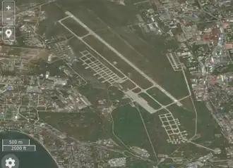 Photographie satellite d'une base aérienne avec une piste au centre de l'image. Le tour de la base est composée de zones urbanisées.