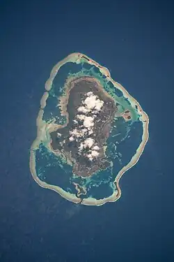île brune et blanche au centre d'un lagon formé par une barrière de corail circulaire, le tout entouré de mer bleue