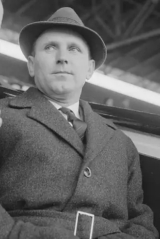 Sur une photographie en noir et blanc, un homme est en costume avec un chapeau, vu de face.