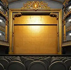 Un grand rideau doré ferme la scène. On distingue les fauteuils du parterre et les balcons.