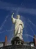 Statue blanche, sur fond de ciel bleu, encadrée par des jets d'eau