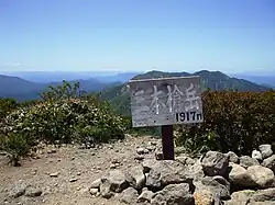 Photo couleur montrant une pancarte, plantée dans un sol gris pierreux partiellement recouvert de buissons verts, en bois annonçant l'altitude d'un sommet de montagne, une chaîne de montagne boisée au second plan et un ciel bleu en arrière-plan.