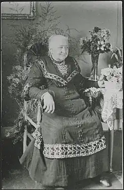 Une dame âgée, assise dans un fauteuil, entourée de fleurs et de feuilles.