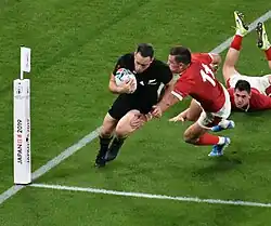 Joueur de face, le ballon sous son bras droit, évitant un adversaire, un autre adversaire, étendu au sol.