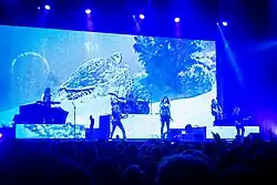 Six membres d'un groupe de rock jouant devant un écran présentant une tortue marine devant un public.