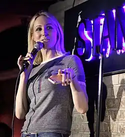 Nikki Glaser tenant un micro, debout devant un mur en brique