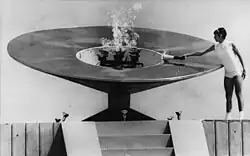 Photo en noir et blanc d'une femme vêtue de blanc portant une torche enflammée, pointant le centre d'une grande vasque en métal.