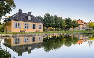  Bâtiment sans étage d'allure classique au bord d'un étang rectiligne. Quelques bancs, collés au bâtiment, au bord de l'étang.