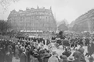 Photo en noir et blanc d'un cortège funéraire (civils et militaires) accompagnant un véhicule, dans une avenue aux immeubles haussmanniens