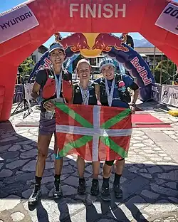 Trois femme tenant un drapeau basque devant une arche d'arrivée rouge.
