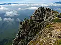 Photo couleur d'une falaise rocheuse. L'arrière-plan est composé d'une nappe de nuages au-dessus d'un massif montagneux boisé.