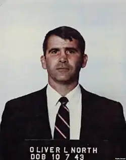 Photographie en couleurs et en buste d'un homme derrière une plaque de détention et portant un costume