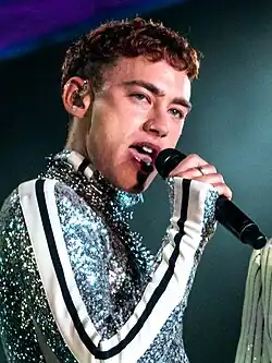 Jeune chanteur mince, rasé de près et aux cheveux courts, il est habillé d'une tenue à paillettes scintillantes et tient très près de sa bouche un microphone en regardant l'horizon.