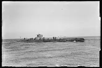 Photo noir et blanc d'un sous-marin sur une mer calme, portant l'inscription « K XII » à la proue, avec une grosse vingtaine d'hommes sur le pont et le kiosque.