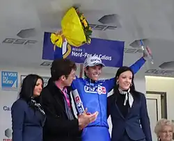 De trois-quarts, quatre personnes sont alignées. Un homme en bleu soulève un bouquet et un trophée.