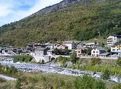 Photographie en couleurs d'un village au bas d'une montagne, le long d'un cours d'eau quasiment à sec.