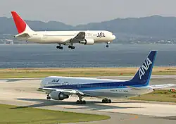 Un appareil de la Japan Airlines, de couleur blanche avec la dérive rouge, au-dessus de la piste avec le train d'atterrissage sorti ; un avion fr All Nippon Airlines dans une livrée bleue et blanche est au roulage.