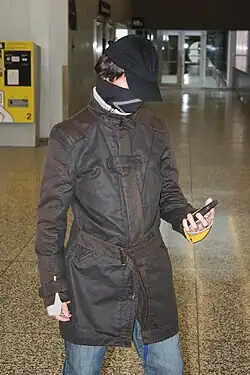 Homme caucasien portant une casquette noire, un foulard noir, un manteau marron, et un jean bleu regardant un téléphone portable avec sa main gauche.