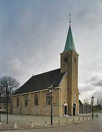 Photographie en couleurs et de trois quarts d'un édifice en briques et de son parvis longé par une rue.