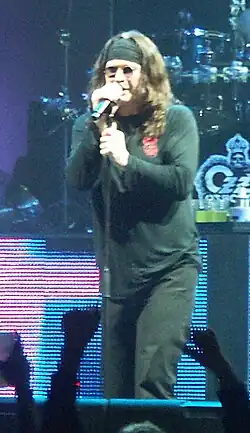 Homme caucasien sur scène habillé de noir et tenant un micro.