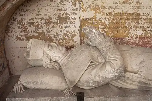 Gisant de pierre représentant un homme, en costume ecclésiastique, joignant les mains.