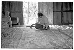 Photo en noir et blanc d'une femme assise en arrière plan dans une grande pièce. Sur le sol s'étend un très grand tissu rempli de motifs géométriques. Une autre femme, assise sur le côté, la regarde.